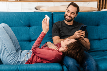 Happy young couple using mobile phones while resting on couch / Photodune, Misc