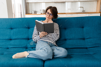 Brooding nice woman in eyeglasses reading book while sitting on couch / Photodune, Misc