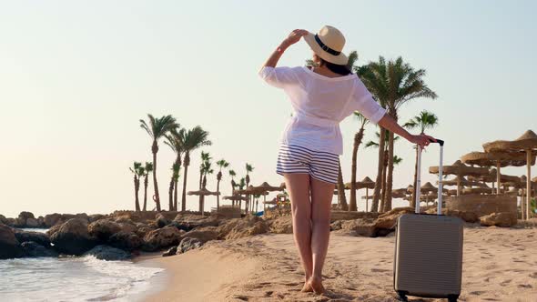 Back View of Young Woman in Summer Clothes and Sun Hat, Holding Travel Bag, Looking at the Sea