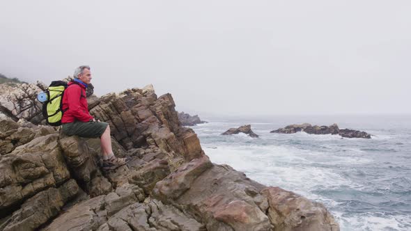 Senior hiker man with backpack sitting on the rocks while hiking near sea shore.