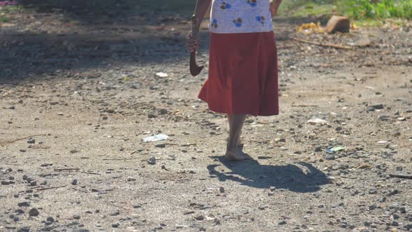 Senior Woman in Skirt with Machete Walks Along Dirty Road