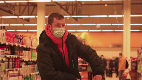 A Young Man Stands with a Trolley in a Supermarket with a Mask on His Face