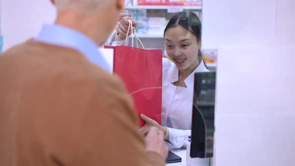 Portrait of Asian Woman Passing Shopping Bag with Drugs to Client in Pharmacy Smiling
