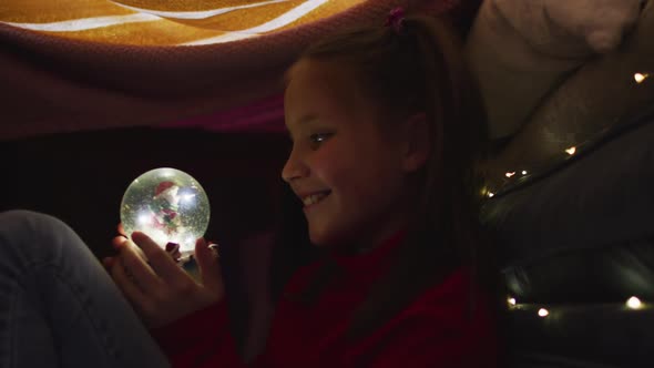 Caucasian girl smiling while holding snow globe under blanket fort during christmas at home