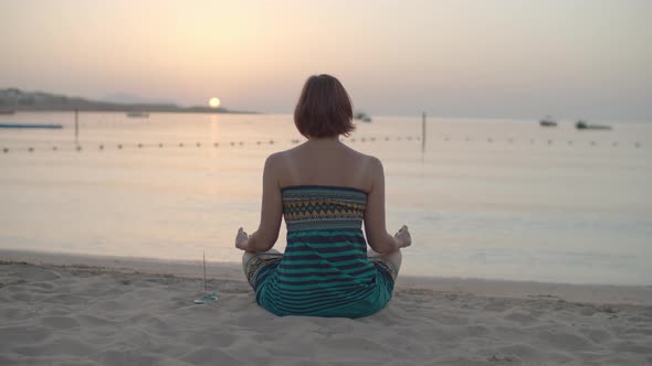 30s Brunette Woman in Dress Sitting in Lotus Position and Meditating on the Beach at Sea Shore