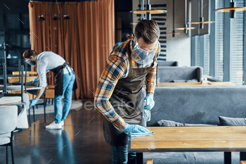 Busy young male waiters in protective workwear spraying disinfectant on tables / Photodune, Misc