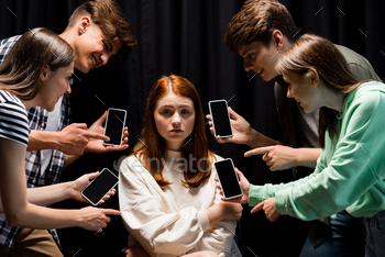 smiling teenagers pointing with fingers at girl during bullying and holding smartphones with blank / Photodune, Misc
