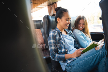 laughing multiethnic female friends reading book during trip on travel bus / Photodune, Misc