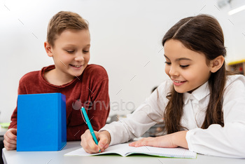 Smiling schoolgirl talking with friend while writing in notebook in classroom / Photodune, Misc