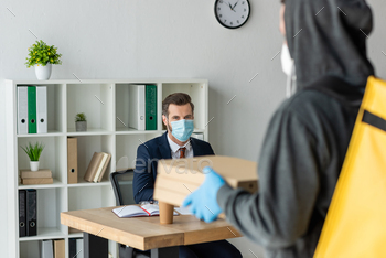 selective focus of food delivery man holding pizza boxes near businessman in medical mask sitting at / Photodune, Misc