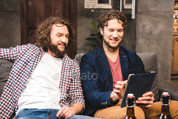 Smiling male friends sitting on sofa and using digital tablet / Photodune, Misc