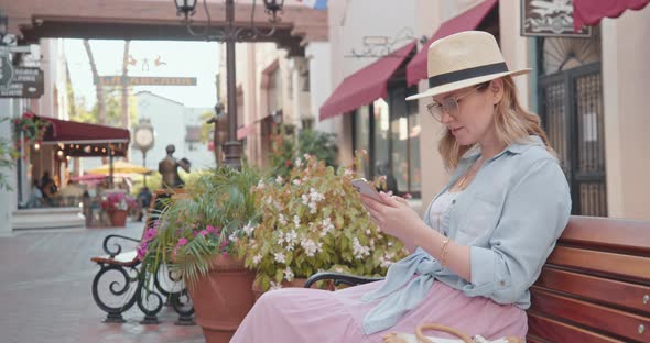 Pretty Woman Is Checking Phone on Summer, Sitting on Bench in Cinematic Old Town