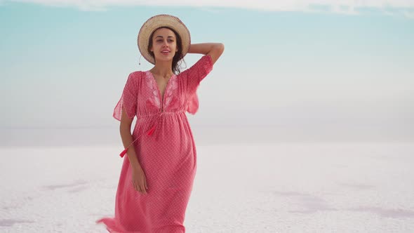 Joyful Freedom Woman in Blowing Pink Dress and Hat Standing on White Endless Salty Beach Like Desert