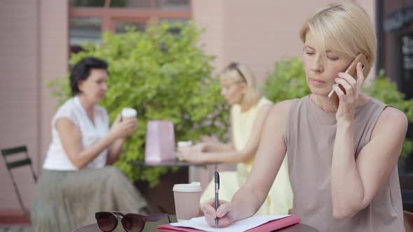 Serious Mid-adult Businesswoman Talking on the Phone and Taking Notes. Portrait of Confident Female