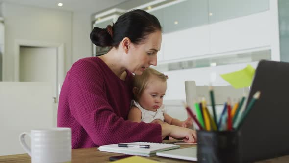 Caucasian mother holding her baby using laptop while working from home