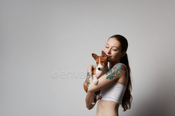 彼女の小さな子犬バセンジー犬を抱きしめてキスするかわいい入れ墨の若い女性の肖像画。 / Photodune, Misc