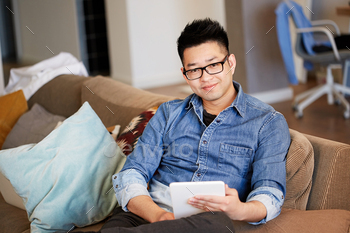 Young Asian man using his electronic tablet while relaxing on comfort sofa at coworking studio / Photodune, Misc