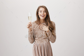  in evening dress smiling and pointing at herself while standing with champagne over gray background. Author shocked she is nominated on award.