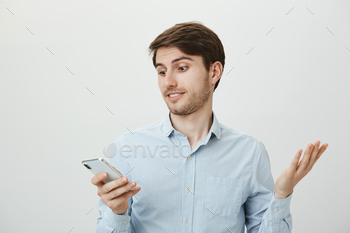 s positive handsome guy in blue shirt shrugging, raising palm in confusion and looking at screen of smartphone, receiving strange reply under post.