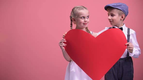Couple of Romantic Kids Hiding Behind Red Heart Cutout and Kissing, First Love