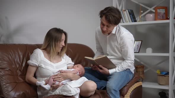 Portrait of Smiling Father Singing Lullaby Reading Book Sitting with Wife and Newborn Son on Couch