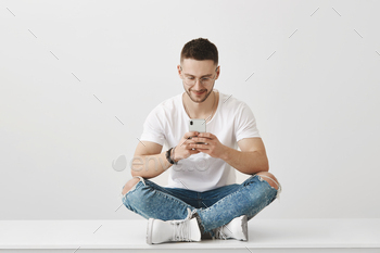 ttractive young coworker sitting in trendy outfit on floor with crossed legs, holding smartphone and looking at screen with excited expression.