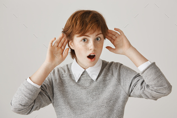 . Portrait of interested shocked redhead woman holding hands near ears while gossiping with friend or overhearing conversation over gray background.
