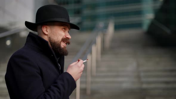 a Mustachioed and Bearded Stylish Man in a Hat and a Dark Coat Smokes a Cigarette on the Stairs Out