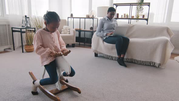 Little Afro Girl Daughter Child Rides Toy Horse Swinging Playing Game Alone While African Black Mom