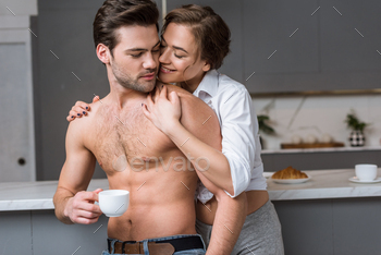 attractive woman hugging shirtless man with cup of coffee in kitchen / Photodune, Misc