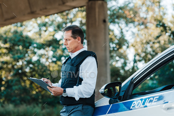 side view of middle aged male police officer in bulletproof vest writing in clipboard near car at / Photodune, Misc