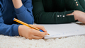Closeup of little boy writing with pen in copybook while lying on carpet in living room. Concept of - PhotoDune Item for Sale