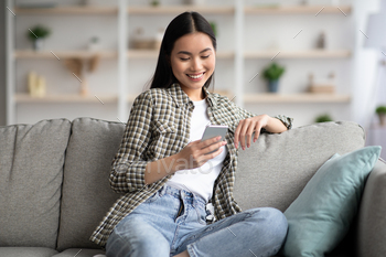 Cheerful asian lady sitting on sofa and using smartphone / Photodune, Misc