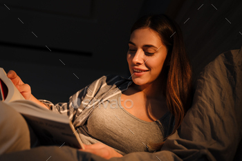 Charming happy woman smiling and reading book while lying in bed / Photodune, Misc