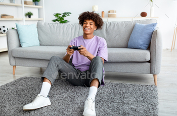 tion, having fun at home, full length. Joyful African American adolescent gamer with joystick sitting on floor, playing computer arcade