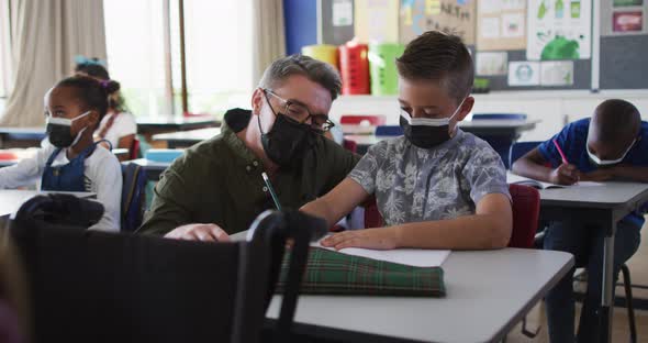 Diverse male teacher helping schoolboy sitting in classroom, all wearing face masks