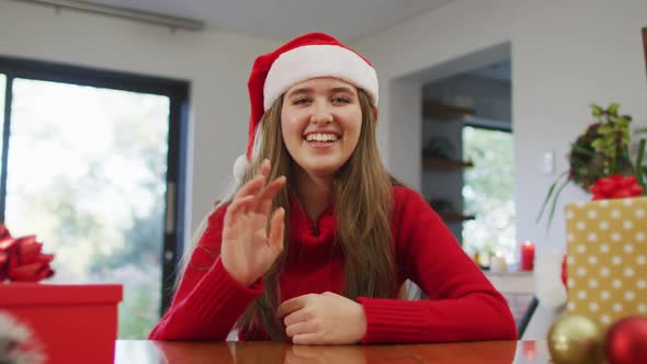 Smiling caucasian woman wearing santa hat making video call at homeh waving