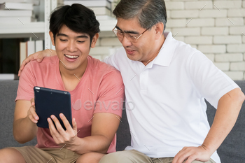 Happy young and senior man using tablet while sitting together on a sofa in a living room. / Photodune, Misc