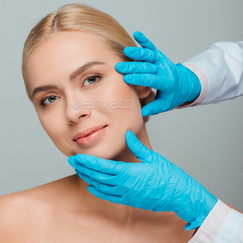 cropped view of beautician in latex gloves touching face of young woman isolated on grey / Photodune, Misc