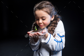 Cute little girl in astronaut costume holding fresh plant in soil on black / Photodune, Misc