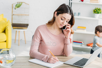 beautiful woman talking on smartphone while sitting at table with laptop at home / Photodune, Misc