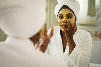 African american woman in bathroom wearing towel on head, looking in mirror and applying face mask / Photodune, Misc
