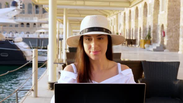 Young Woman in Summer Clothes and Sun Hat, Uses Laptop, Sitting Alone at Table on Promenade By the
