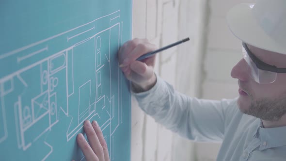 Young engineer-architect in a bright room in a helmet and glasses works with blueprints 