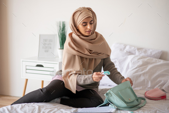 Smart girl sitting on the bed packing some beauty products to bag