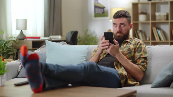 Young Man Browsing News in Mobile Phone While Sitting on Sofa in His Living Room