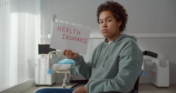 Young African Patient in Wheelchair at Hospital Ward Holding Health Insurance Poster