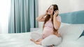 Happy smiling pregnant woman in pajamas listening to music with headphones on bed at morning - PhotoDune Item for Sale