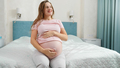 Portrait of happy smiling pregnant woman in pajamas sitting on bed at sunny morning - PhotoDune Item for Sale