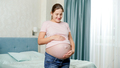Smiling pregnant woman caressing and touching big belly while standing in bedroom with big window - PhotoDune Item for Sale